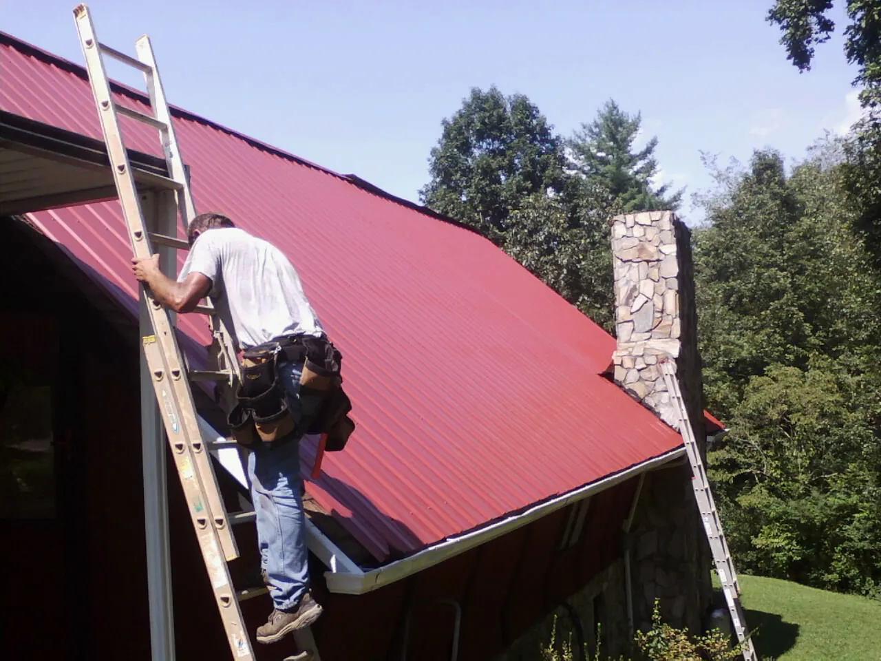 Roofer on a ladder beside a red metal roof and stone chimney — Soffit & Fascia Repair by Roofing Brothers in Spring Glen